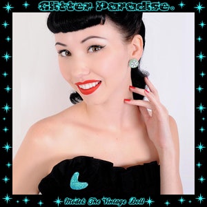 May include: A woman with dark hair and red lipstick is wearing a black strapless dress and a pair of turquoise and silver earrings. The earrings are round and have a floral design. The woman is smiling and looking at the camera. The text "Glitter Paradise" is at the top of the image.