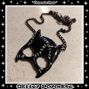 May include: Black catwoman mask necklace with a black chain. The mask has white stitching details. The necklace is on a white glitter background.