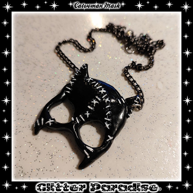 May include: Black catwoman mask necklace with a black chain. The mask has white stitching details. The necklace is on a white glitter background.