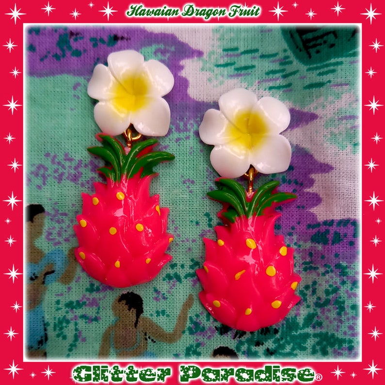 May include: A pair of dangle earrings featuring a pink dragon fruit with yellow dots and a white plumeria flower. The earrings are on a green and white fabric background.