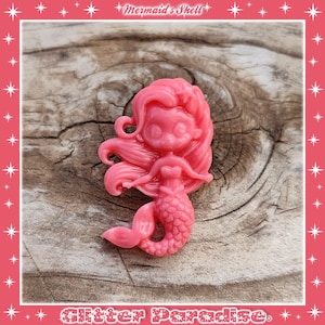 Puede incluir: Una figura de sirena rosa con cabello suelto y una cola escamosa. La figura es de un color sólido y tiene un diseño de dibujos animados. La imagen tiene el texto "Mermaid's Shell" y "Glitter Paradise."