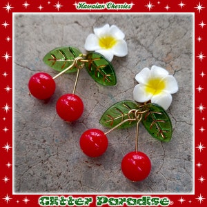 May include: A pair of dangle earrings featuring red cherry charms, green leaves, and white plumeria flowers. The earrings are labeled "Hawaiian Cherries" and "Glitter Paradise".