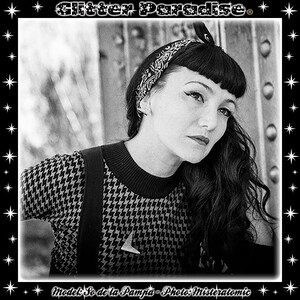 May include: Black and white photo of a woman with dark hair wearing a black and white patterned sweater, a black bandana, and a pair of suspenders. The photo is framed with a black border and stars.