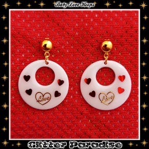 May include: White hoop earrings with gold-coloured details. Each earring has a circular design with red and black heart-shaped decorations and the word "Love" in gold script. The earrings are suspended from a gold-coloured ball.