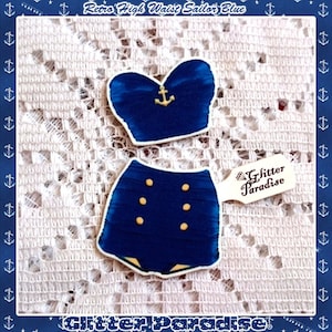 May include: A blue and gold sailor-themed swimsuit with a high-waisted bottom and a strapless top. The top has a gold anchor on the front. The bottom has four gold buttons on the front. The swimsuit is on a white lace background. "Glitter Paradise" is written on a white tag.