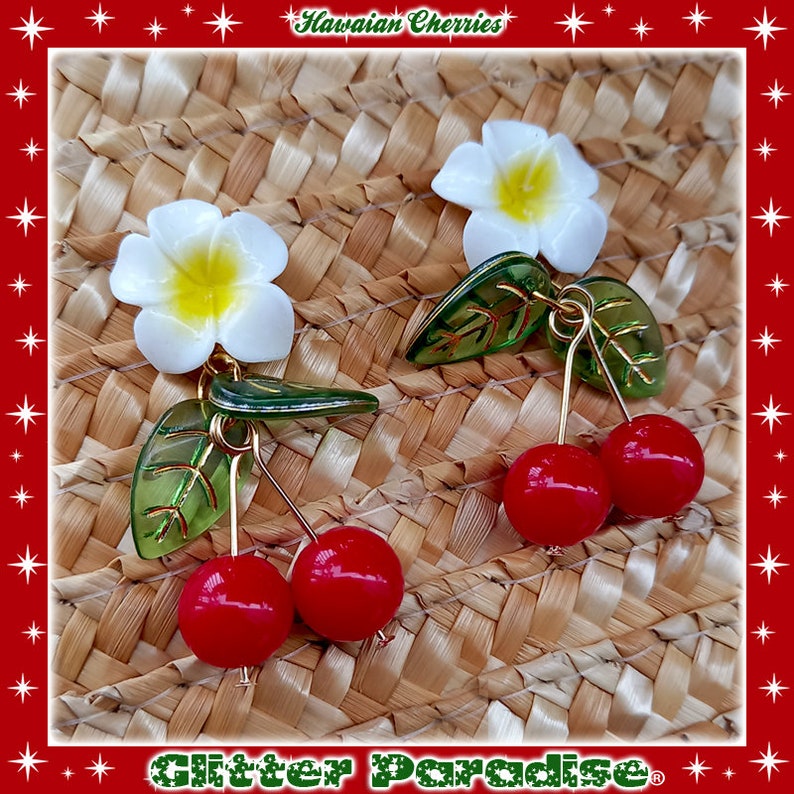 May include: A pair of earrings featuring a white plumeria flower and two red cherries with green leaves. The earrings are on a woven straw background. The text "Hawaiian Cherries" and "Glitter Paradise" are visible in the image.