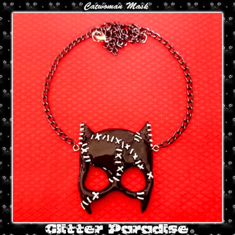 May include: Black catwoman mask necklace with a silver chain. The mask has white stitching details and is on a red background. The text "Glitter Paradise" is at the bottom of the image.