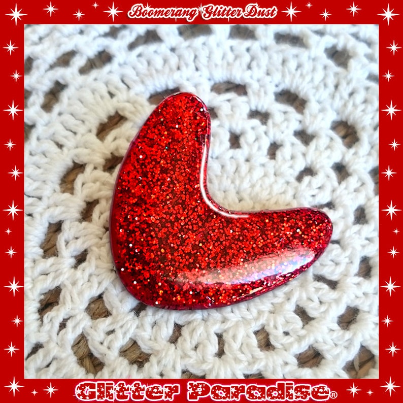 May include: Red glitter boomerang shaped resin brooch. The brooch is on a white crocheted doily.