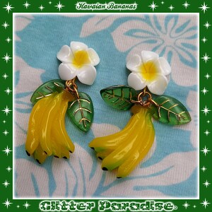 May include: A pair of dangle earrings featuring a cluster of yellow bananas with green leaves and a white plumeria flower. The earrings are labeled "Hawaiian Bananas".