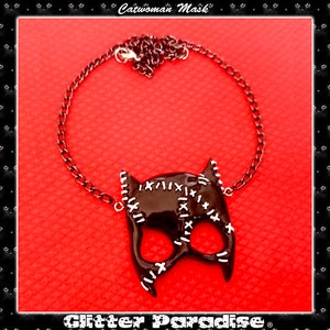 May include: A black catwoman mask necklace with a silver chain. The mask has white stitching details and a glossy finish. The necklace is on a red background.