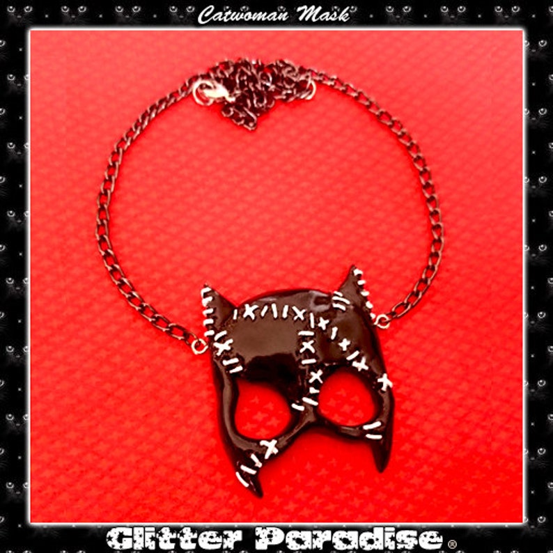 May include: A black catwoman mask necklace with a silver chain. The mask has white stitching details and a glossy finish. The necklace is on a red background.