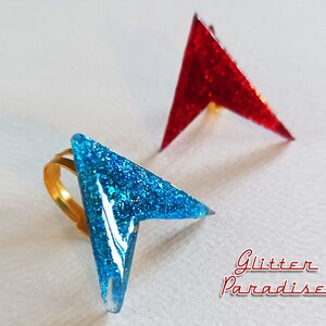 May include: Two arrow-shaped rings with glitter embedded in the resin. The ring on the left is blue and has a gold band. The ring on the right is red.
