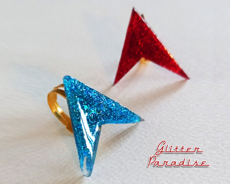 May include: Two arrow-shaped rings with glitter embedded in the resin. The ring on the left is blue and has a gold band. The ring on the right is red.