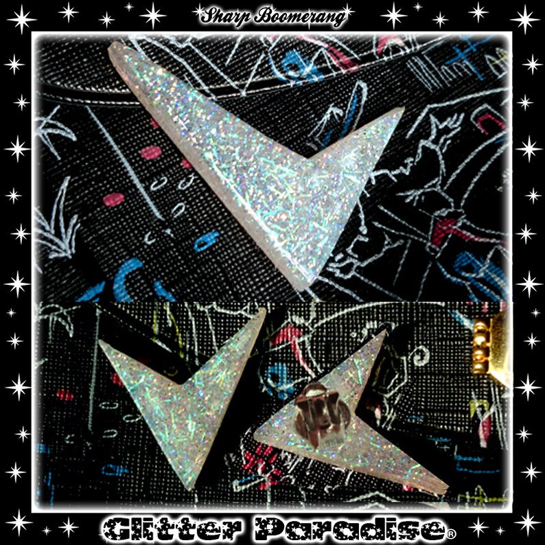 May include: Two silver glitter boomerang earrings with a sharp point. The earrings are on a black fabric background with a white and blue pattern.