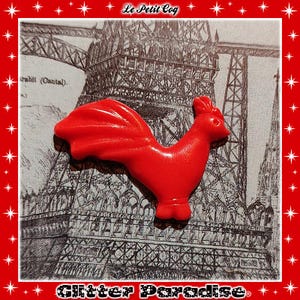 May include: A vibrant red rooster ornament, set against a black and white Eiffel Tower illustration. The image has a red border with white stars and the text "Le Petit Coq" and "Glitter Paradise".