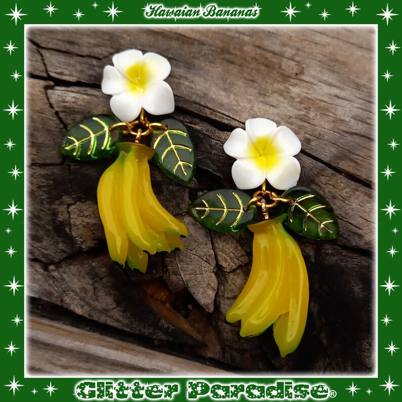 May include: A pair of earrings featuring a cluster of yellow bananas with a white plumeria flower and green leaves. The earrings are made of polymer clay and have a gold finish. The earrings are labeled "Hawaiian Bananas".