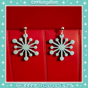 Może przedstawiać: Para jasnoniebieskich kolczyków w kształcie płatków śniegu ze złotymi sztyftami. Kolczyki są prezentowane na czerwonym tle. Tekst "Mid-Century Burst" i "Glitter Paradise" są widoczne.