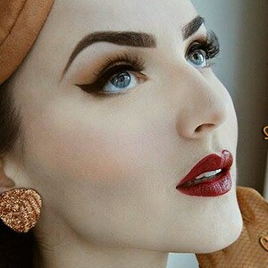 May include: A woman with dark brown hair and a brown hat is wearing dramatic eye makeup with winged eyeliner and red lipstick. The text "Glitter Paradise" is visible in the background.