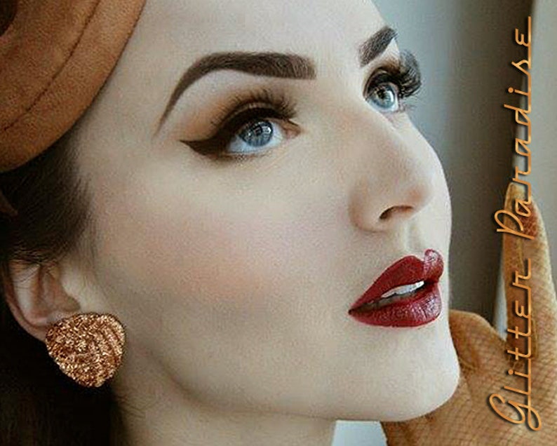 May include: A woman with dark brown hair and a brown hat is wearing dramatic eye makeup with winged eyeliner and red lipstick. The text "Glitter Paradise" is visible in the background.