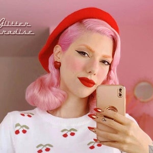 May include: A woman with pink hair and red lipstick is wearing a red beret, a white sweater with cherry embroidery, and red heart-shaped earrings. She is holding a gold smartphone and taking a selfie. The background is pink.