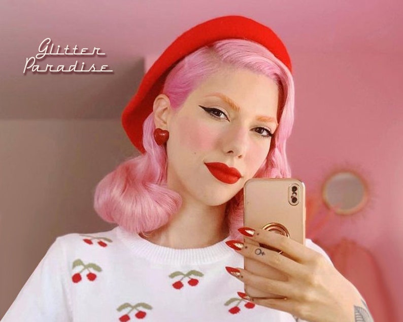 May include: A woman with pink hair and red lipstick is wearing a red beret, a white sweater with cherry embroidery, and red heart-shaped earrings. She is holding a gold smartphone and taking a selfie. The background is pink.