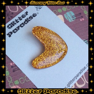 May include: Gold glitter boomerang shaped resin brooch with the text "Glitter Paradise" on the back.