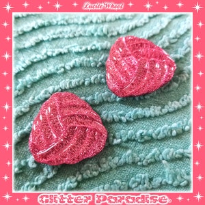 May include: Two pink glitter heart-shaped buttons with a textured design. The buttons are on a light blue textured fabric.