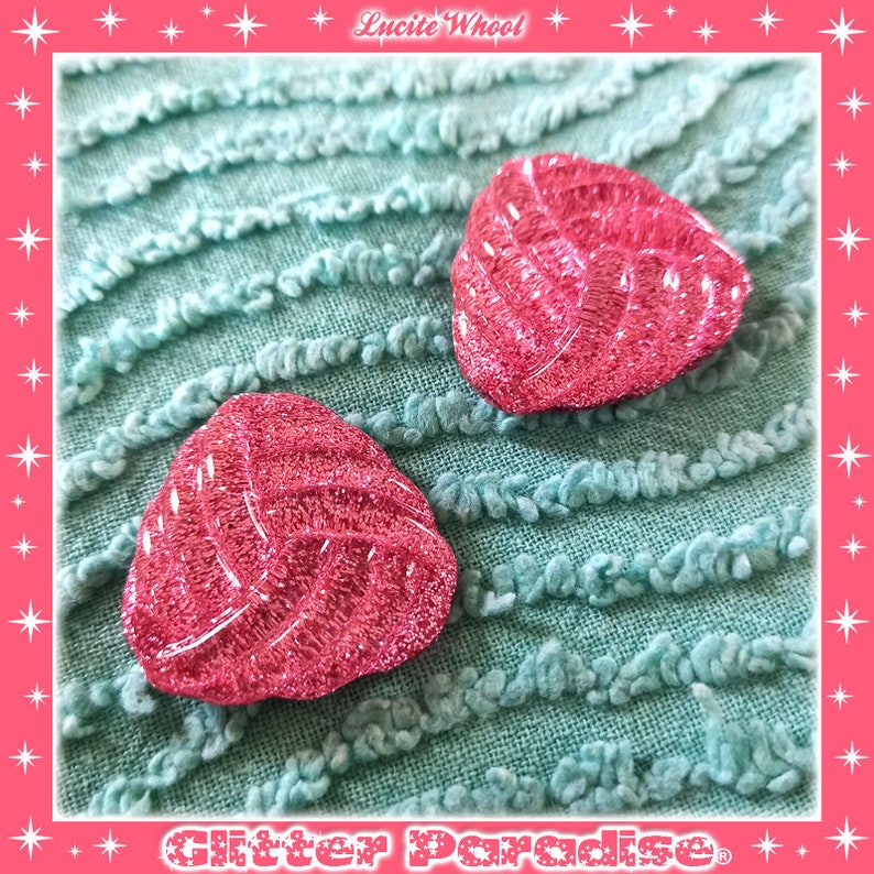 May include: Two pink glitter heart-shaped buttons with a textured design. The buttons are on a light blue textured fabric.