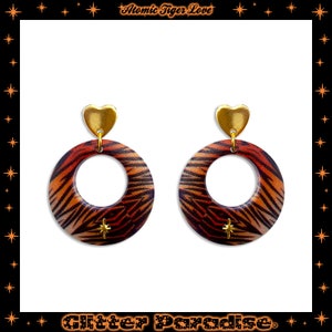 Pode incluir: Um par de brincos de coração dourado com círculos de estampa de tigre. Os brincos são feitos de madeira e têm um acabamento dourado.