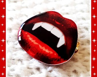 Könnte beinhalten: Eine glänzende Brosche in Form von geöffneten Lippen mit spitzen weißen Fangzähnen. Die Lippen sind in einem tiefen, leuchtenden Rot mit dunklen Schattierungen gehalten. Der Text Vampire Kiss steht oben und Glitter Paradise unten im Bild.