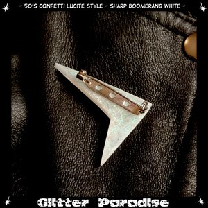May include: A white boomerang-shaped brooch with a silver pin and glitter embedded in the lucite. The brooch is on a black leather background.