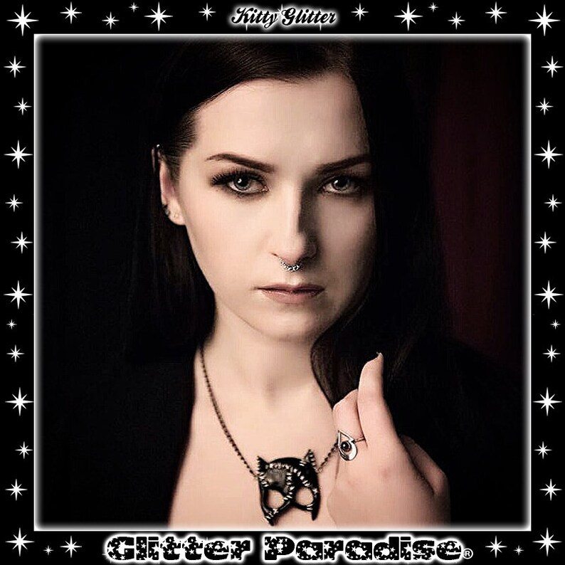 May include: A woman with dark hair and dark eye makeup is wearing a black necklace with a cat-shaped pendant. She is also wearing a silver ring with a teardrop-shaped stone. The background is dark and blurry.