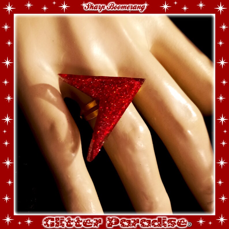 May include: A red glitter ring shaped like a sharp boomerang. The ring is on a finger of a hand.