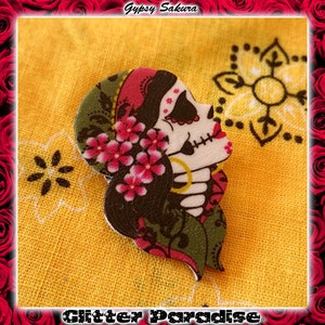 Peut inclure: Une découpe en bois peinte à la main, colorée, d'une femme avec un visage de crâne de sucre, portant un foulard rouge et des embellissements floraux. La découpe est sur un fond de tissu jaune avec un motif blanc et noir.