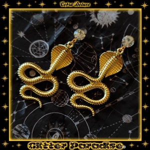 Puede incluir: Pendientes de cobra dorados con detalles de cristal de roca transparente. Los pendientes están sobre un fondo negro con un patrón de estrellas.