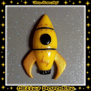 Può includere: Una statuetta a forma di razzo gialla con finitura lucida, caratterizzata da un oblò circolare nero, fasce orizzontali nere e un emblema a forma di fulmine dorato sul corpo. Il razzo ha due pinne curve glitterate dorate e un ugello nero.