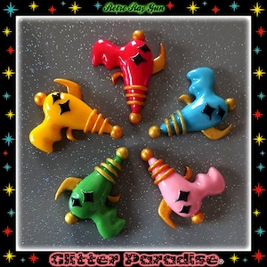 May include: Five colorful toy ray guns with gold accents and black star details. The guns are arranged in a circle on a gray glittery background. The text "Retro Ray Gun" is at the top of the image and "Glitter Paradise" is at the bottom.