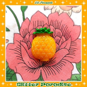 May include: A small, orange pineapple-shaped charm with a green leafy top. The charm is set against a backdrop of a large pink flower on a white and pink patterned fabric. The text "Lil Pineapple" and "Glitter Paradise" are in the image.