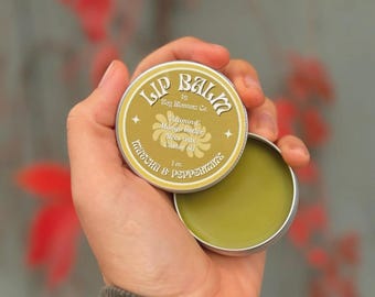 Könnte beinhalten: Eine runde, silberne Lippenbalsam-Dose mit den Worten "Lip Balm" in einer dekorativen Schriftart. Der Deckel ist geöffnet und zeigt einen grünen Balsam. Das Etikett listet auch die Inhaltsstoffe auf: Vitamin & Mango butter, Bees wax, Castor oil, und Kakuna & Peppermints.