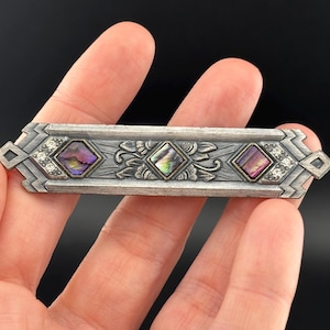 May include: A silver bar pin with three iridescent, square-cut stones. The pin features floral and geometric designs, with small clear stones set on each end. The pin is held in a hand against a dark background.