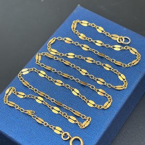 May include: A gold chain necklace featuring a series of elongated, flattened oval links connected by small circular rings. The chain has a polished finish and includes a circular spring ring clasp at one end, showcasing a consistent, repeating pattern.