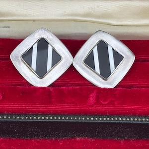 May include: A pair of silver cufflinks with a geometric design. Each cufflink has a black and white striped square within a diamond-shaped frame. The cufflinks are presented in a red velvet lined box.