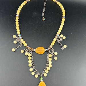 May include: A beaded necklace featuring round yellow beads, metallic chains, and two leaf-shaped yellow pendants. The design includes small iridescent crystals, silver-toned metal links, and a single green bead accent above the bottom pendant.