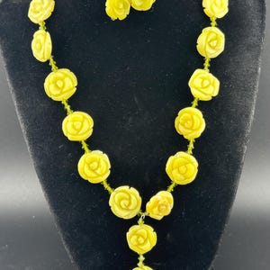 May include: A yellow rose necklace and matching earrings set. The necklace features carved rose pendants strung on a green beaded chain. The earrings have a similar rose design. The jewelry is displayed on a black mannequin.