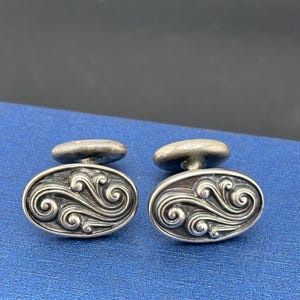 May include: A pair of oval-shaped silver cufflinks featuring a raised, swirling scrollwork pattern with darkened recessed areas. Each cufflink has a smooth, rounded toggle backing attached to the center of the oval face, showcasing metallic luster.