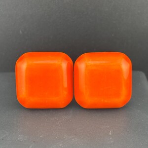 Op de afbeelding: Een paar feloranje vierkante knopen met afgeronde hoeken en een gladde, glanzende afwerking. Elke knoop heeft een plat oppervlak met licht afgeschuinde randen en toont een levendige, effen kleur over de gehele vierkante vorm.