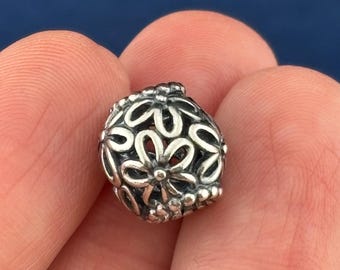 May include: A silver charm bead with a floral design. The bead is spherical, with openwork flower shapes and small beaded accents. The charm is held between fingers against a blue background.