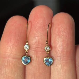 May include: A pair of gold earrings featuring a heart-shaped blue gemstone and a small, round, clear gemstone. The earrings are suspended from a gold hook.
