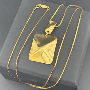 Pode incluir: Um colar dourado com um pingente retangular. O pingente tem uma superfície texturizada e pende de uma corrente delicada. O colar é exibido em uma superfície cinza escuro.