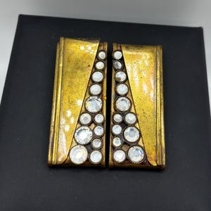 以下が含まれることがあります： ヴィンテージのゴールドトーンのメタルクリップオンイヤリング2つ。各イヤリングは長方形で、透明で輝くラインストーンの三角形が特徴です。イヤリングは黒い背景に配置されています。